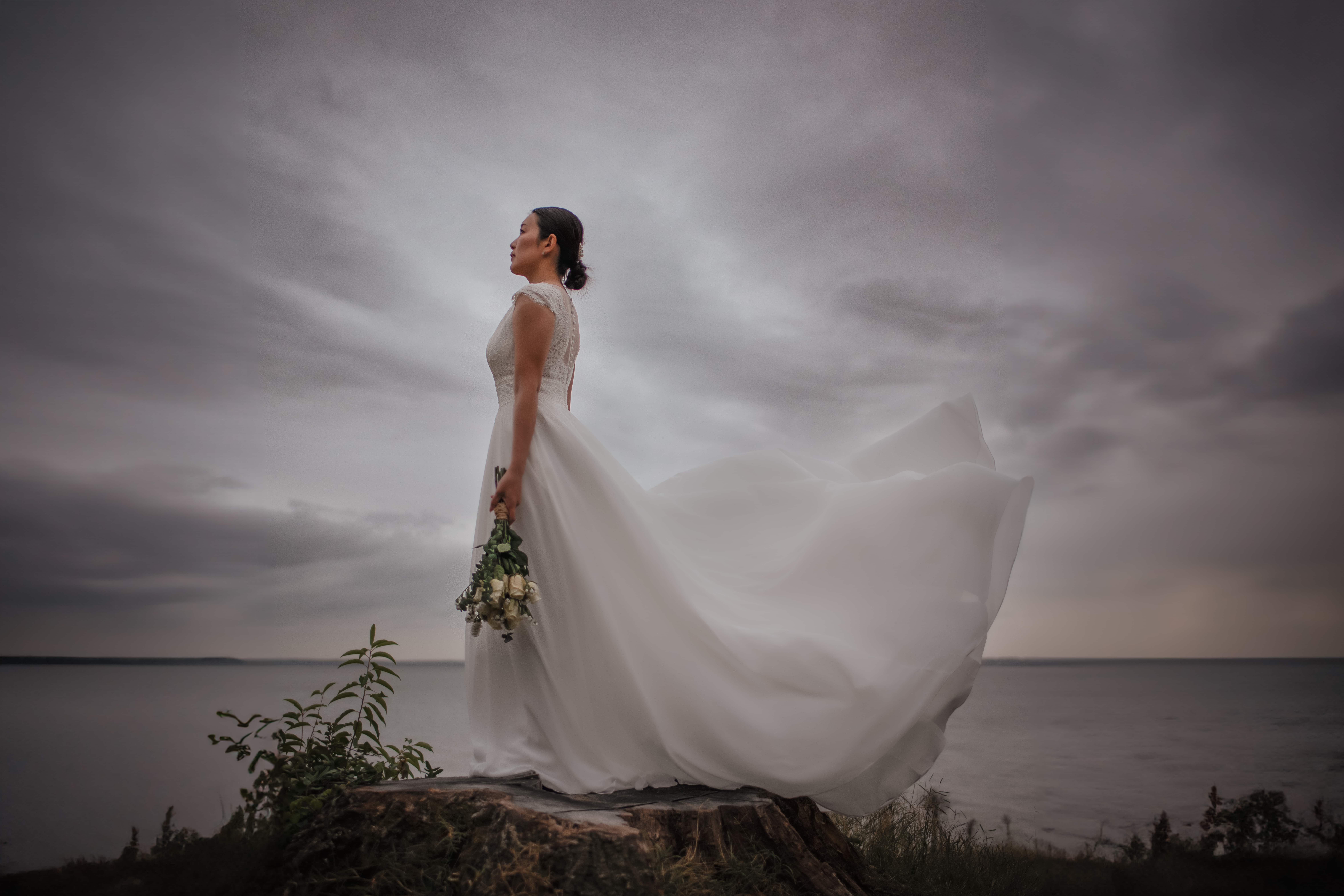 Bride in flowing white wedding dress holding bouquet, standing on a cliffside by the water under dramatic stormy skies